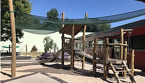 Das Bild zeigt den Außenbereich des Gartens mit einem Klettergerüst, Sandkasten, eines Trampolins und einer Vogelnestschaukel